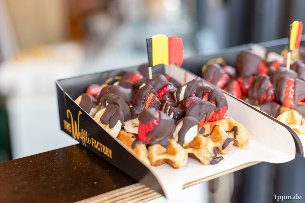 Eine dicke belgische Waffel mit einer kleinen belgischen Flagge. Die Waffel ist reichlich mit Bannenscheinen, Erdbeeren und einer dicken Schokoladenschicht garniert.