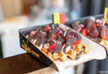 Eine dicke belgische Waffel mit einer kleinen belgischen Flagge. Die Waffel ist reichlich mit Bannenscheinen, Erdbeeren und einer dicken Schokoladenschicht garniert.