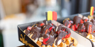 Eine dicke belgische Waffel mit einer kleinen belgischen Flagge. Die Waffel ist reichlich mit Bannenscheinen, Erdbeeren und einer dicken Schokoladenschicht garniert.