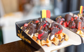 Eine dicke belgische Waffel mit einer kleinen belgischen Flagge. Die Waffel ist reichlich mit Bannenscheinen, Erdbeeren und einer dicken Schokoladenschicht garniert.