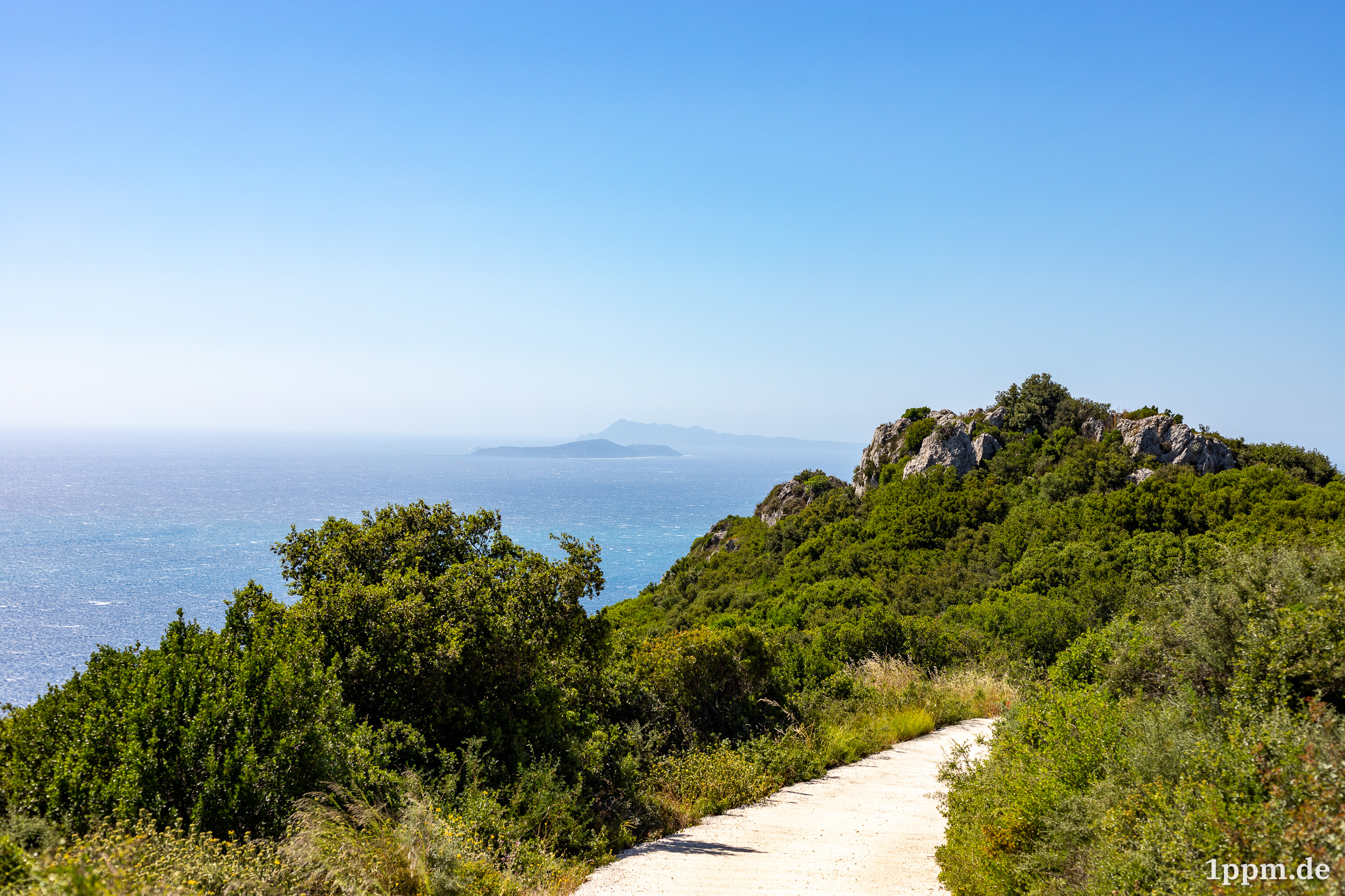 Blick auf das Meer, das ein wenig diesig ist. Im Hintergrund erkennt man zwei Felsformationen im Meer. Im Vordergrund Teile eines Wegs und grüne Büsche.