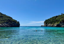 Eine Meerbucht vom Strand aus, klares Wasser, man sieht große Steine am Grund, links und rechts ragen Felsen mit Grün obendrauf aus dem Wasser. Man erahnte einige schwimmende Menschen.