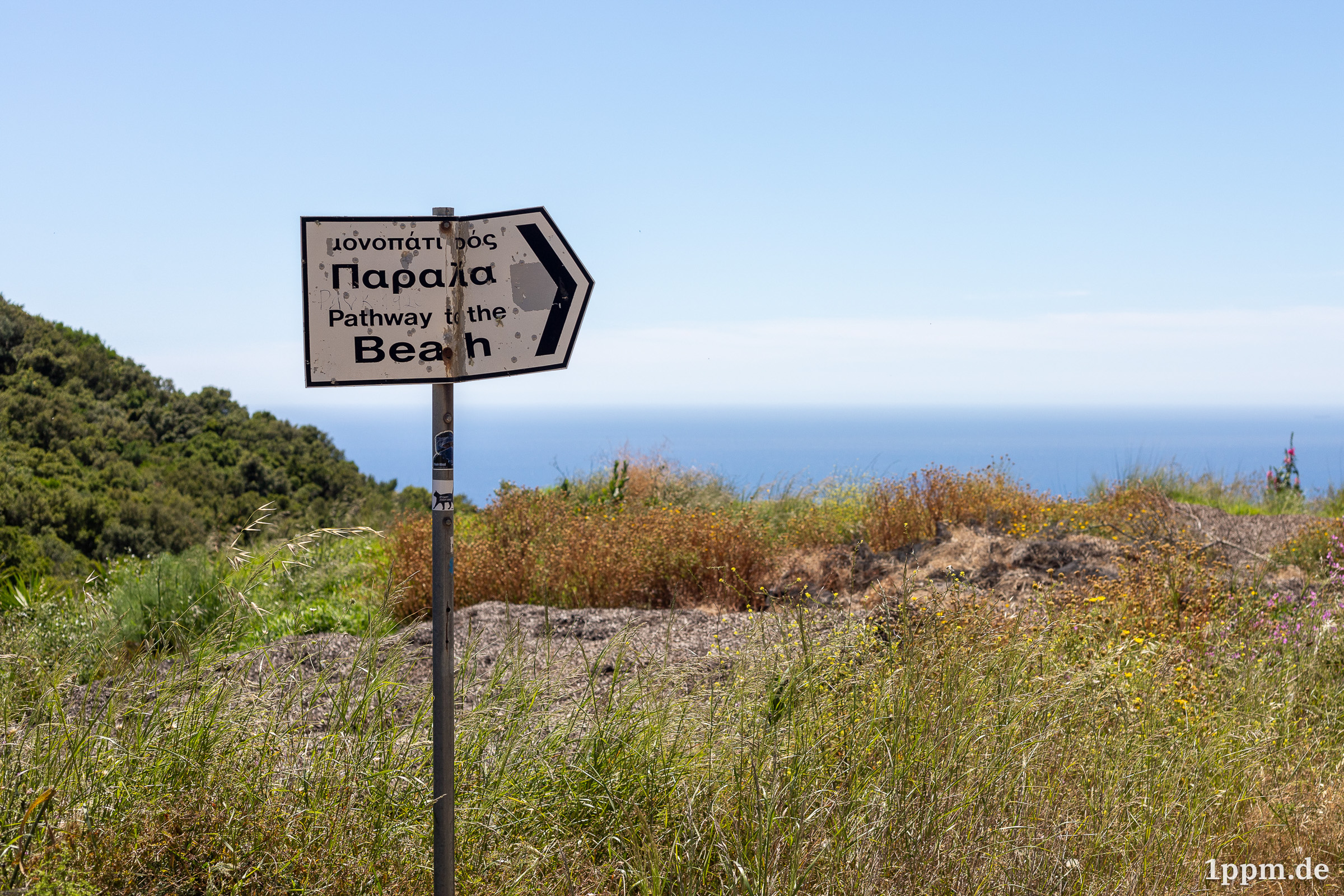 Ein nach rechts weisendes Schild mit einer griechischen Aufschrift und der englischen Übersetzung darunter: "Pathway to the beach". Das Schild ist schon sehr verschlissen, verbogen und verbeult.