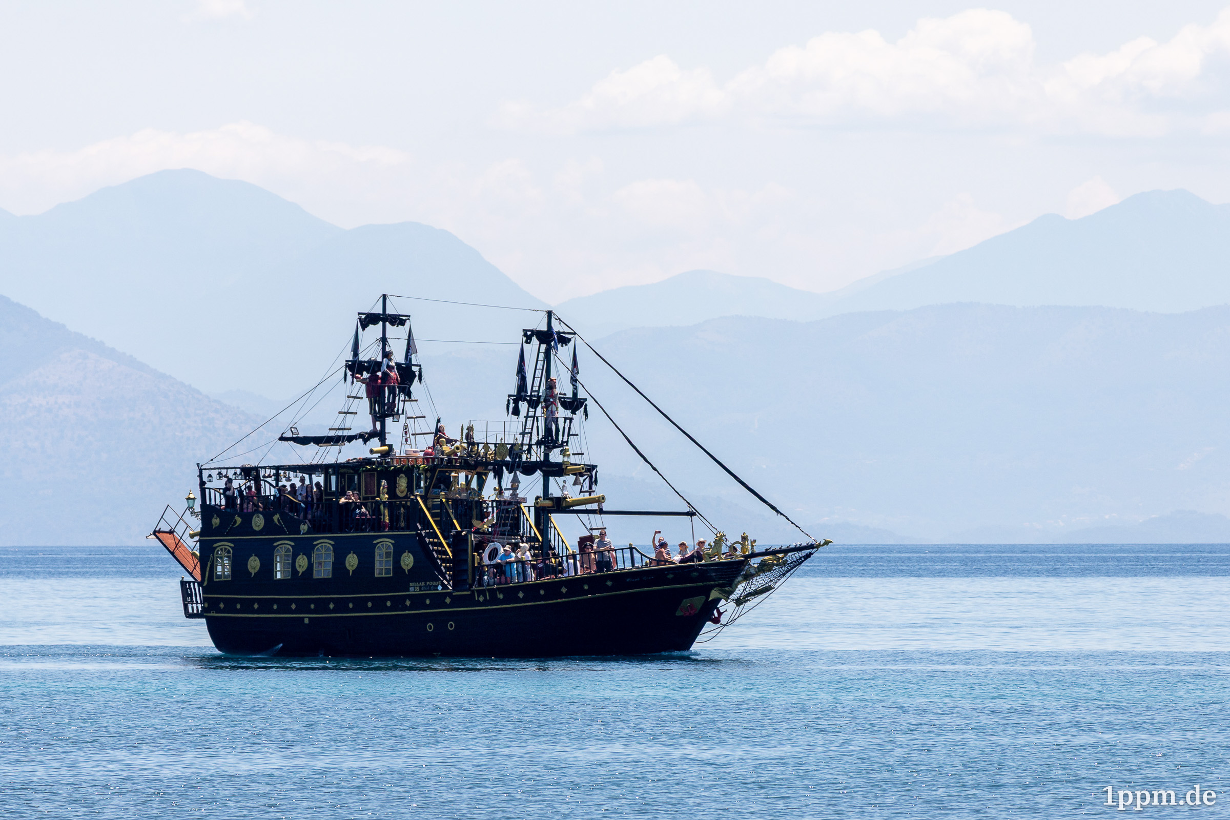 Ein schwarzes Schiff mit goldenen Ornamenten, reich verziert, mit zwei Ausgucken und Piratenflagge. Viele Menschen sind an Bord erkennbar.