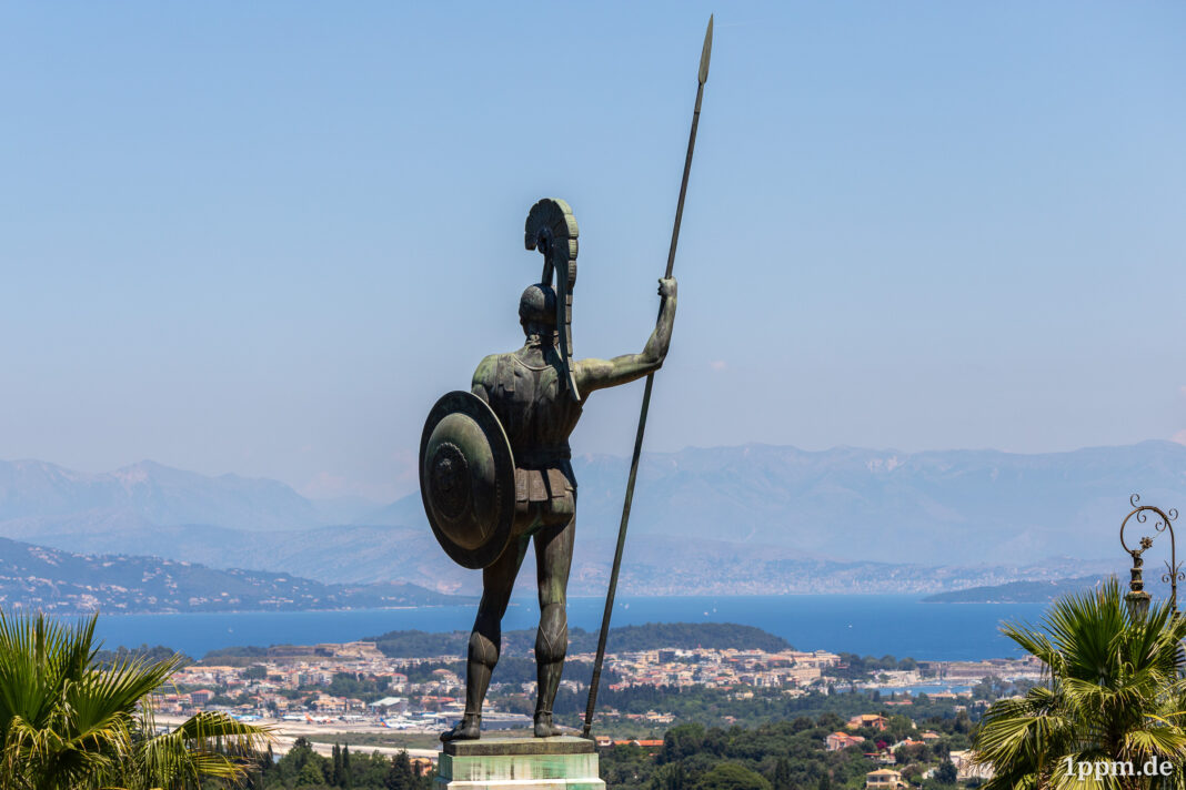 Eine große Statue eines nackten Kriegers mit Schild und Speer von hinten. Er blickt von weit oben auf eine Meeresbucht, etliche Häuser sind zu erkennen.