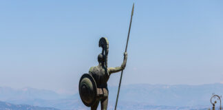 Eine große Statue eines nackten Kriegers mit Schild und Speer von hinten. Er blickt von weit oben auf eine Meeresbucht, etliche Häuser sind zu erkennen.