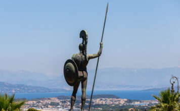 Eine große Statue eines nackten Kriegers mit Schild und Speer von hinten. Er blickt von weit oben auf eine Meeresbucht, etliche Häuser sind zu erkennen.