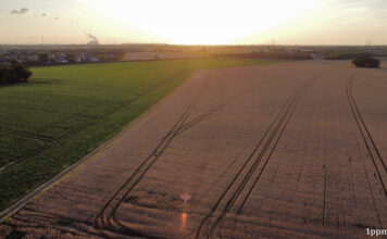 Drohnenaufnahme von einem Feld und einer Wiese daneben, am Horizont ist eine Stadt mit rauchendem Kraftwerk erkennbar. Die Sonne geht gerade unter.