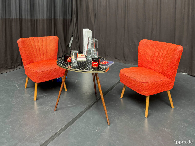 Zwei orangerote Fünfziger-Jahre-Sessel, dazwischen ein Nierentisch mit Tassen, Gläsern und Glaskaraffe darauf. Im Hintergrund noch ein weißer Standventilator.