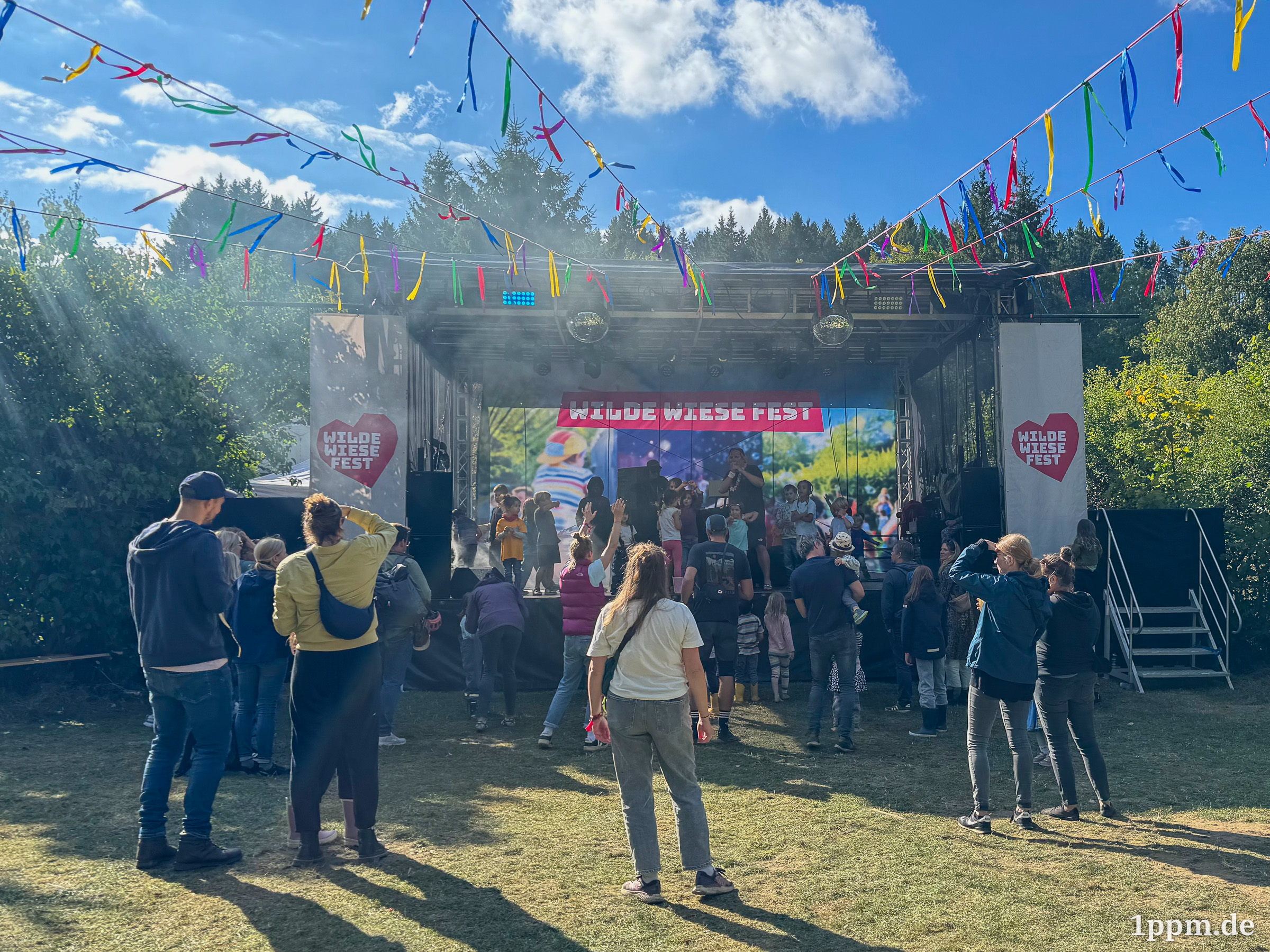 Eine Bühne mit Beschriftungen „Wilde Wiese Fest“, davor einige Erwachsene und Kinder, auf der Bühne stehen ebenfalls eine Menge Kinder.