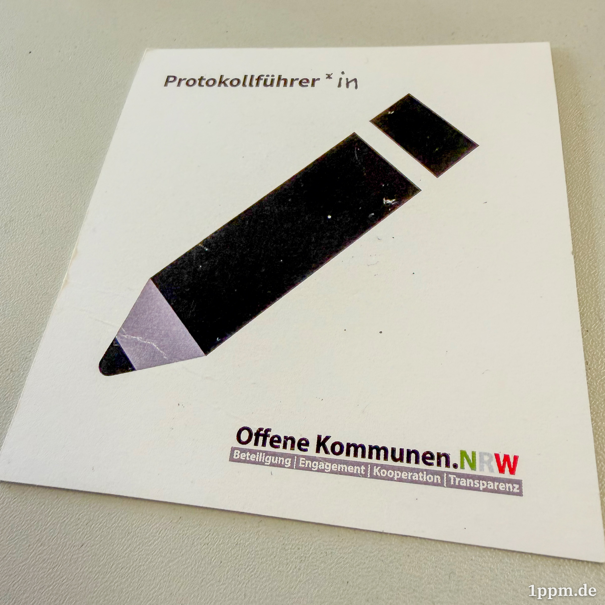 Ein größerer Zettel mit einem großen Stift-Symbol. Oben links steht "Protokollführer*in", unten rechts ist das Logo von "Offene Kommunen NRW".