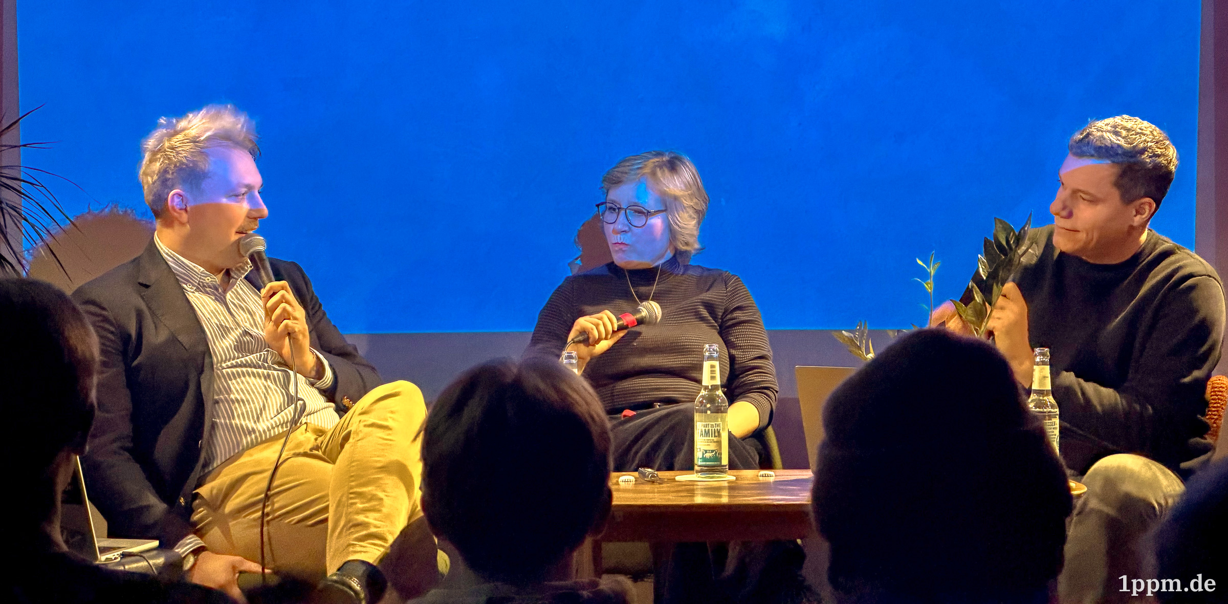 Drei Personen sitzen auf einer Bühne vor blauem Hintergrund, zwei halten Mikrofone, auf dem Tisch stehen Getränke.