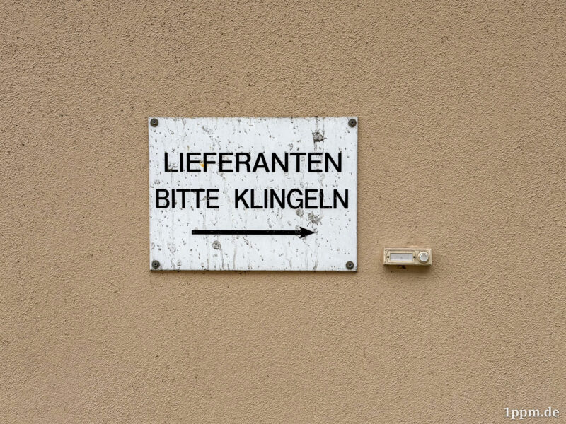 Ein Schild an einer braunen Hauswand: „Lieferanten bitte klingeln“, darunter ein Pfeil, der nach rechts zeigt, wo eine Klingel ist.