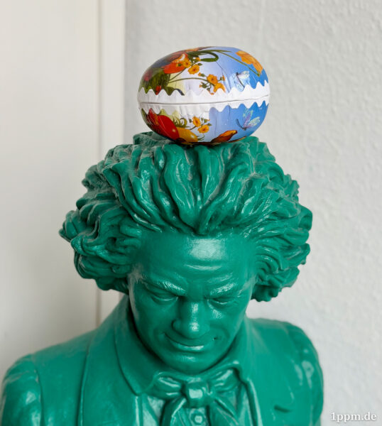 Eine grüne Beethovenfigur (bzw. deren Kopf), auf die ein größeres Papp-Ei abgelegt wurde.
