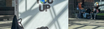 Großes weißes Plakat mit dem Text 'NEVER GONNA GIVE YOU UP' und einer Erde als Buchstabe 'O' in einer Industriehalle mit Personen im Hintergrund