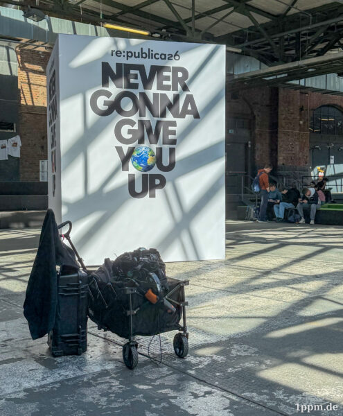 Großes weißes Plakat mit dem Text 'NEVER GONNA GIVE YOU UP' und einer Erde als Buchstabe 'O' in einer Industriehalle mit Personen im Hintergrund