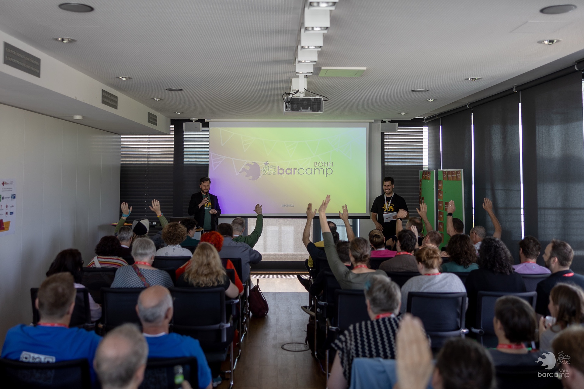 Publikum in einem Konferenzraum hebt die Hände während einer Präsentation mit zwei Sprechern vor einer Leinwand mit dem Schriftzug BarCamp Bonn