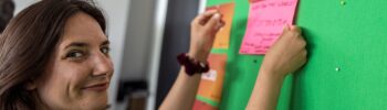 Frau mit dunklen Haaren befestigt einen rosa Zettel an einer grünen Pinnwand mit orangefarbenen Schildern im Hintergrund