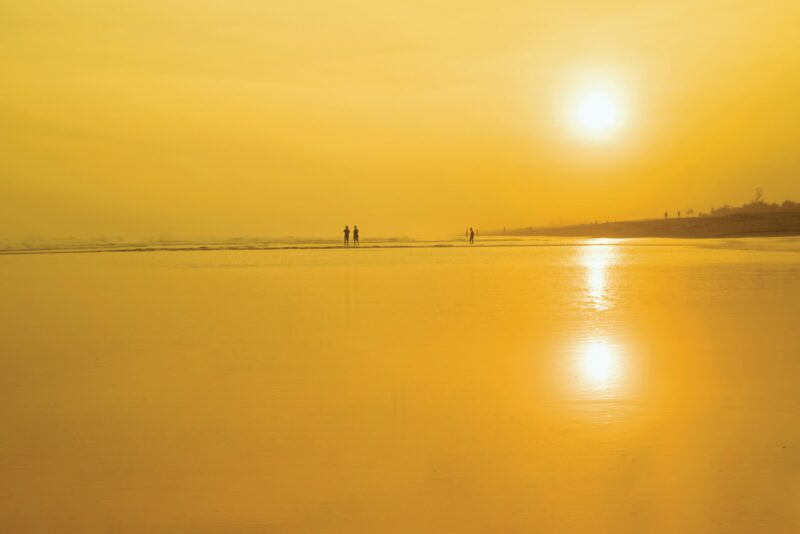Strand mit spiegelndem Wasser und zwei Personen in der Ferne bei Sonnenuntergang