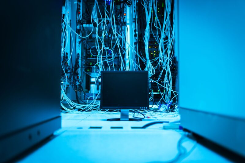 Ein Monitor auf dem Boden, dahinter ein Serverschrank mit unzähligen wirren Kabeln. Das ganze Bild ist in blaues Licht getaucht.
