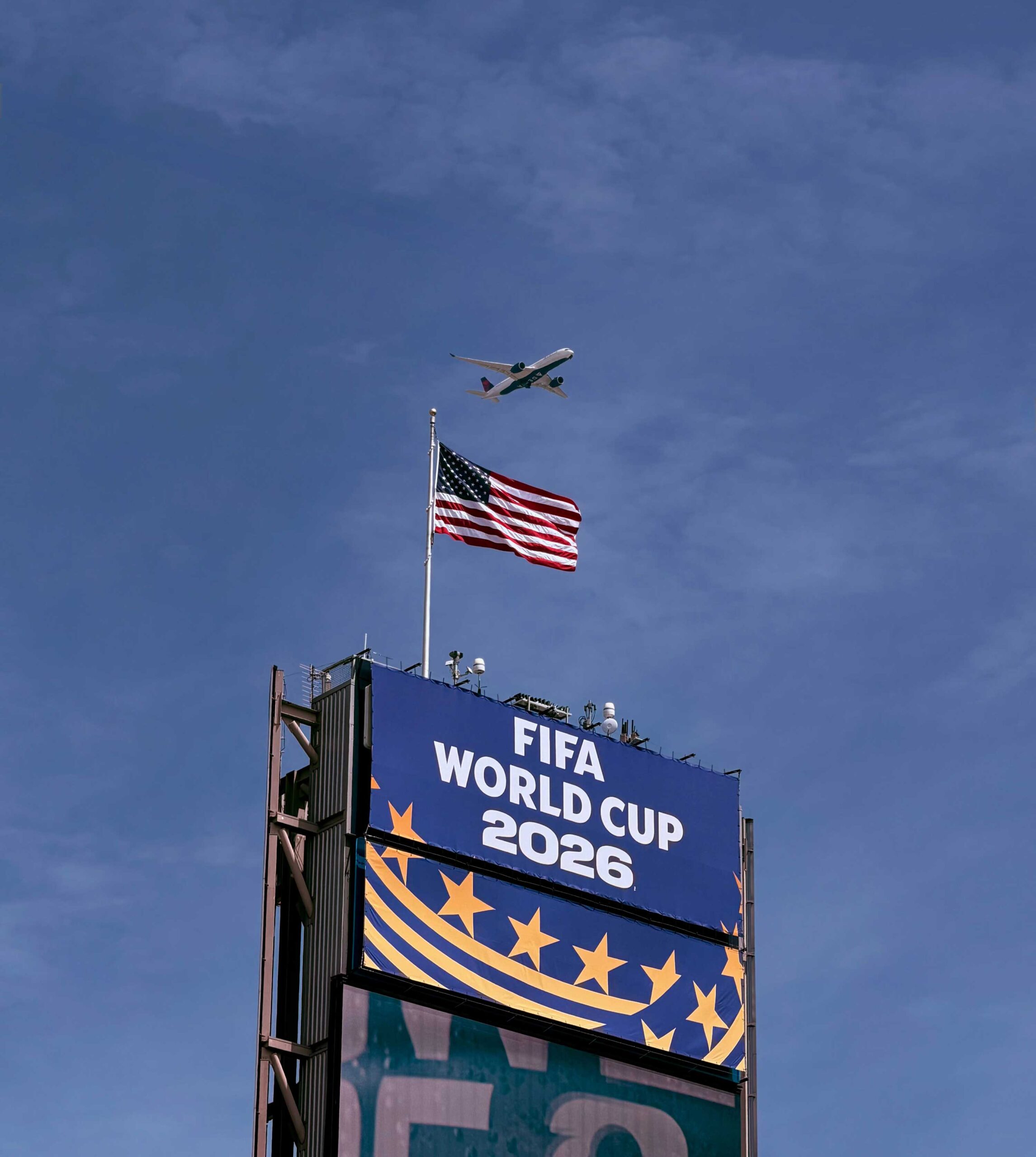 Flugzeug fliegt über einem Schild mit der Aufschrift „FIFA Weltmeisterschaft 2026“ und amerikanischer Flagge, klarer blauer Himmel.