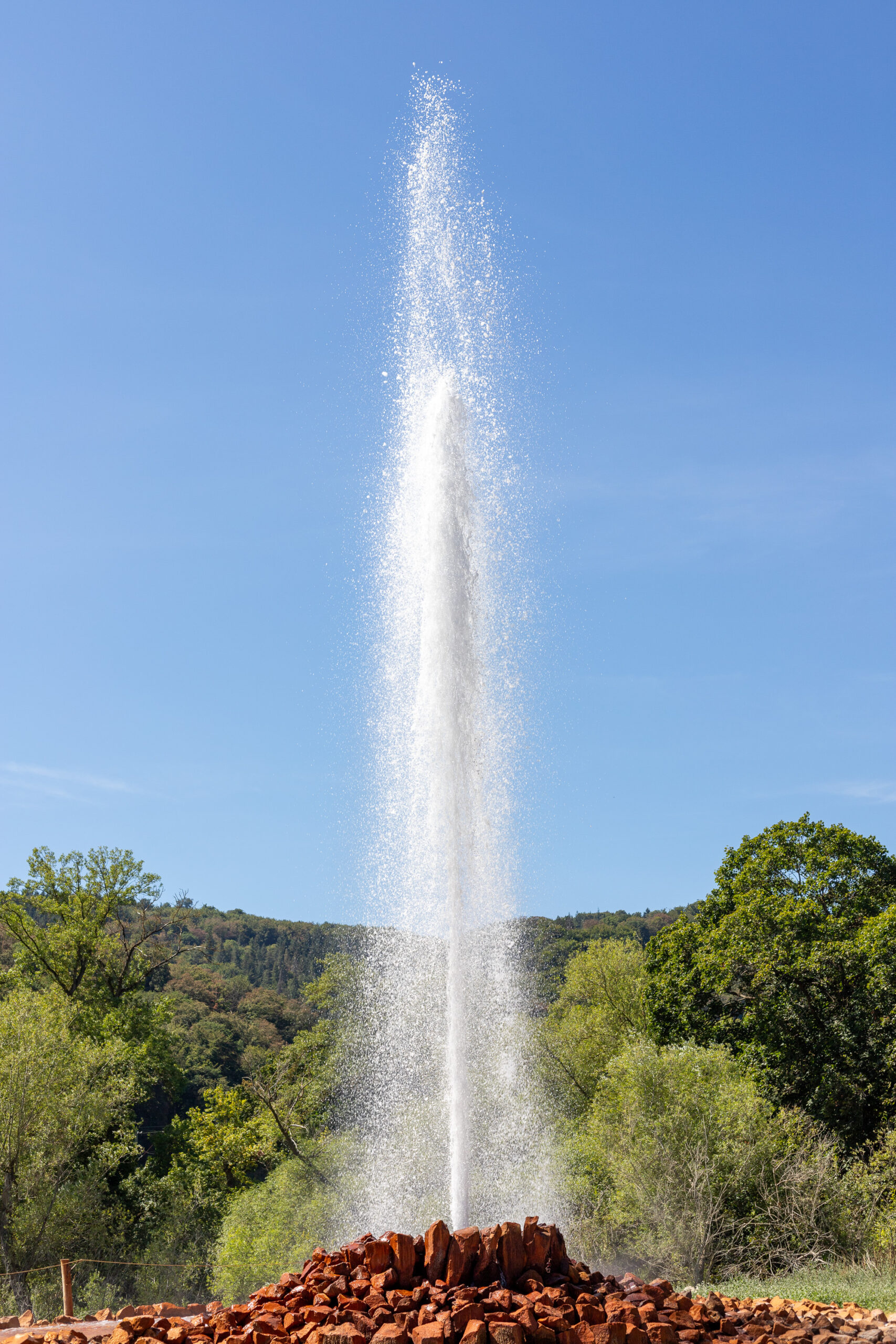 Wasserfontäne, die hoch in den blauen Himmel über einer Landschaft mit Bäumen und Hügeln schießt