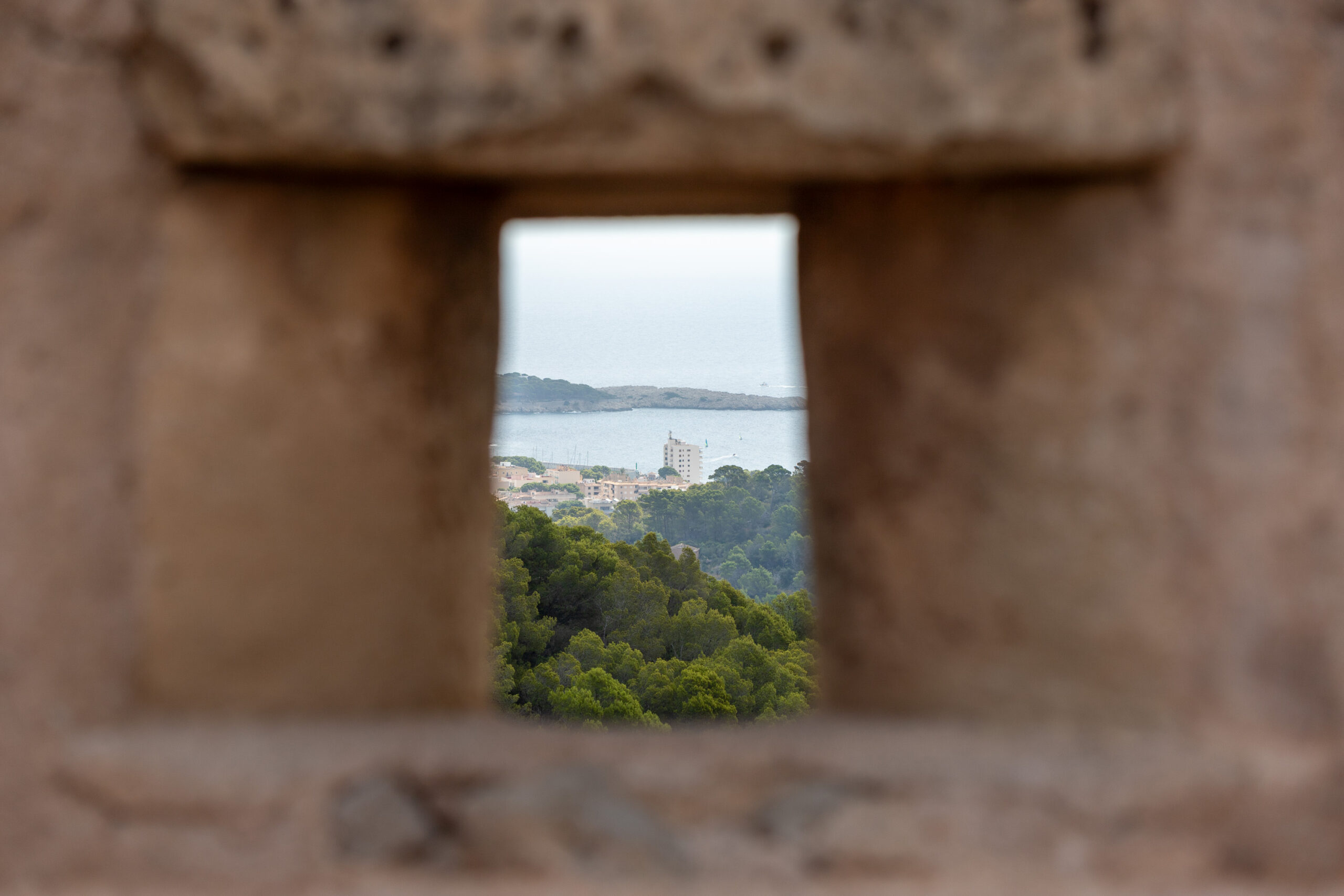 Blick durch ein rechteckiges Mauerfenster auf eine Küstenstadt mit Gebäuden, Bäumen und Wasser im Hintergrund
