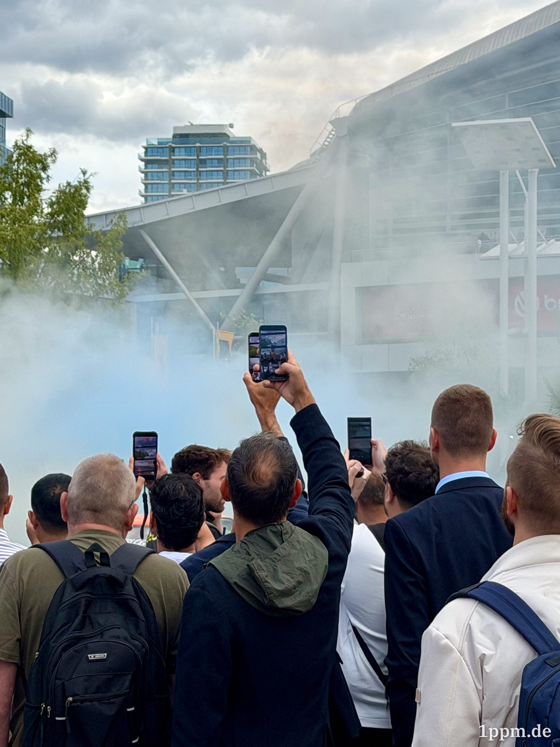 Menschenmenge von hinten vor einem modernen Gebäude, einige halten Smartphones hoch, Rauch oder Nebel liegt in der Luft